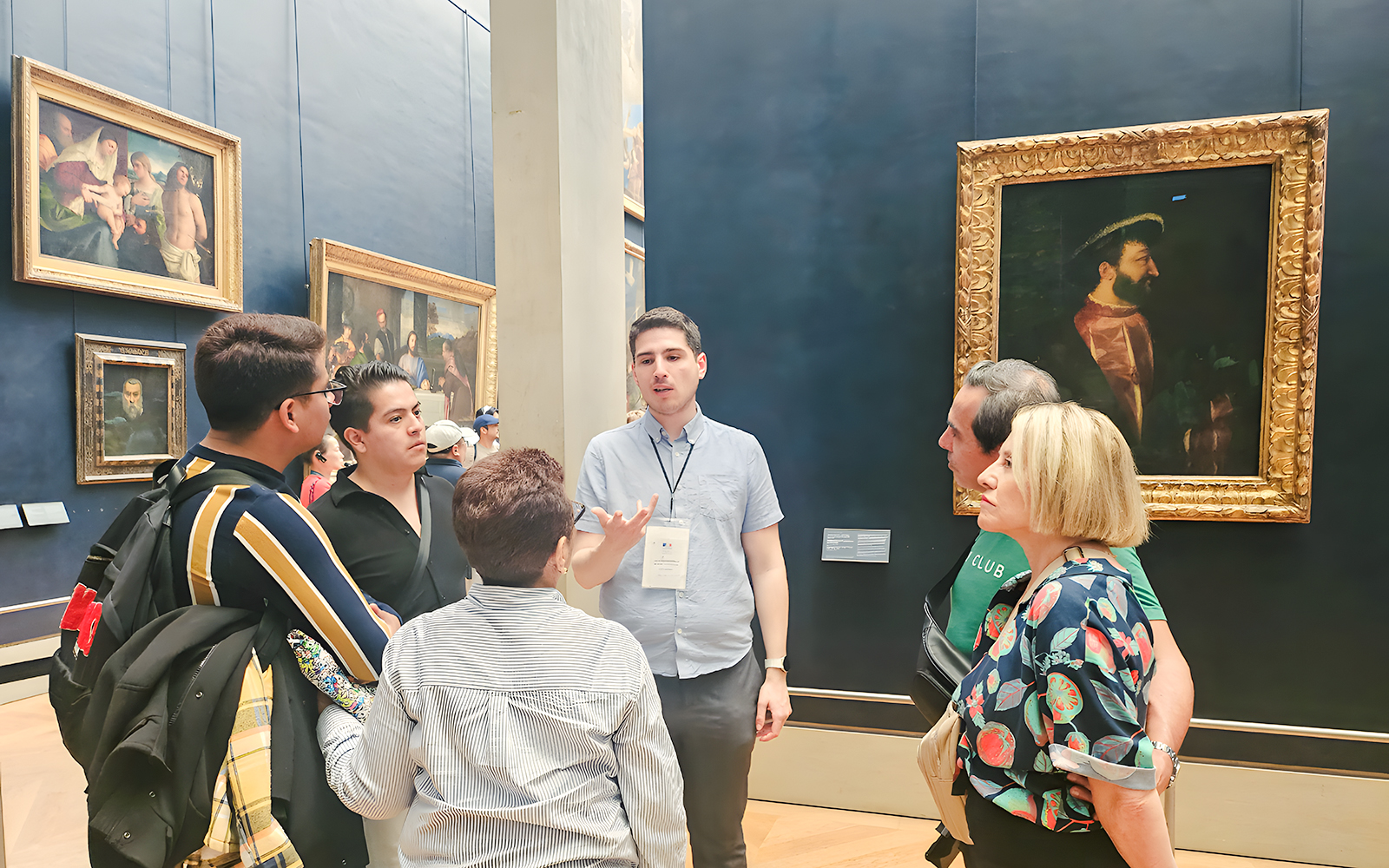Group of people listening to a guide inside the Louvre Museum, Paris, with paintings in the background.