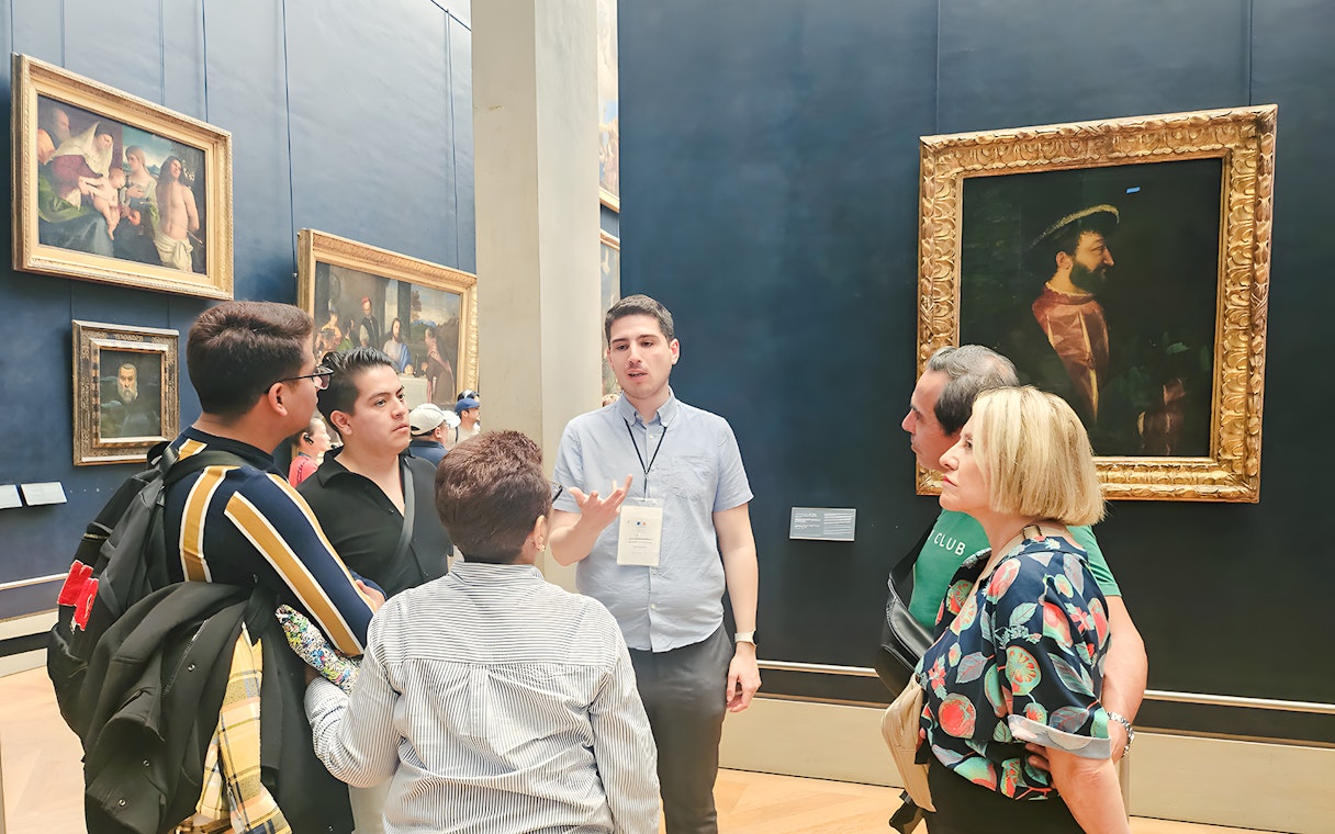 Group of people listening to a guide inside the Louvre Museum, Paris, with paintings in the background.