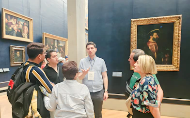 Group of people listening to a guide inside the Louvre Museum, Paris, with paintings in the background.