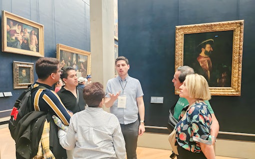 Group of people listening to a guide inside the Louvre Museum, Paris, with paintings in the background.