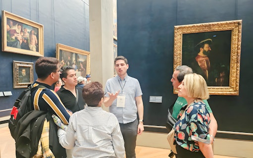 Group of people listening to a guide inside the Louvre Museum, Paris, with paintings in the background.