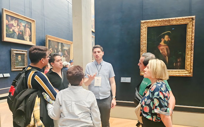 Group of people listening to a guide inside the Louvre Museum, Paris, with paintings in the background.