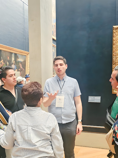 Group of people listening to a guide inside the Louvre Museum, Paris, with paintings in the background.