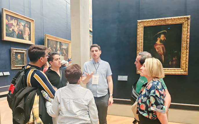Group of people listening to a guide inside the Louvre Museum, Paris, with paintings in the background.