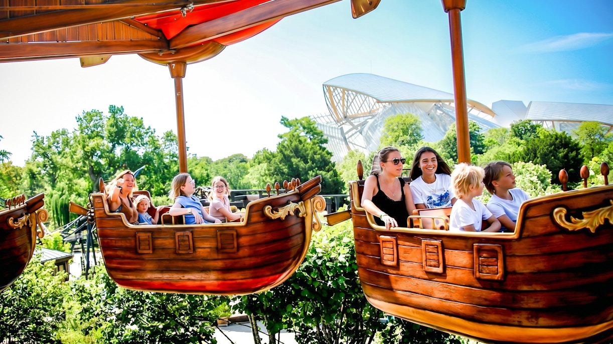 Families enjoying a boat ride at Jardin d'Acclimatation, Paris, with lush greenery and modern architecture in the background.