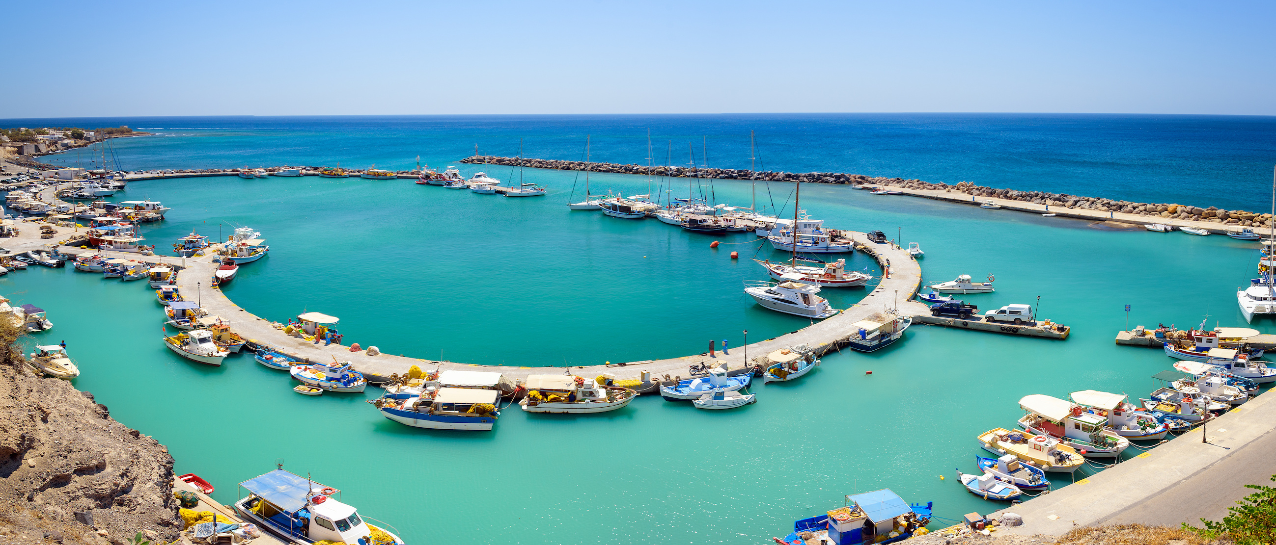 Boats docked at Vlychada port in Santorini, Greece, with clear blue water and a curved pier.