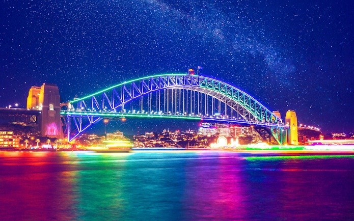 Sydney Harbour Bridge illuminated at night with colorful lights reflecting on the water.