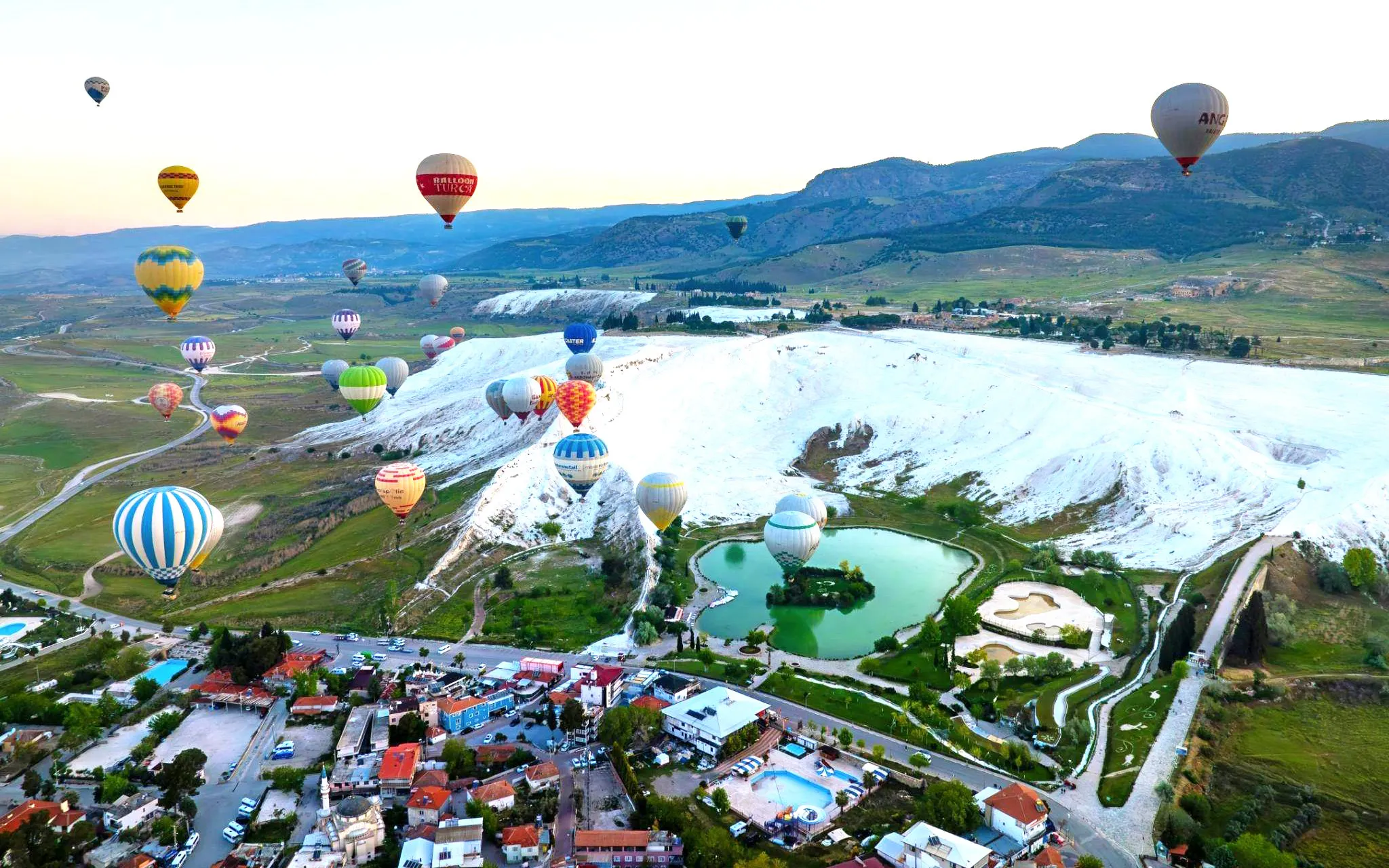 Hot air balloons over Pamukkale's travertine terraces and green landscape.