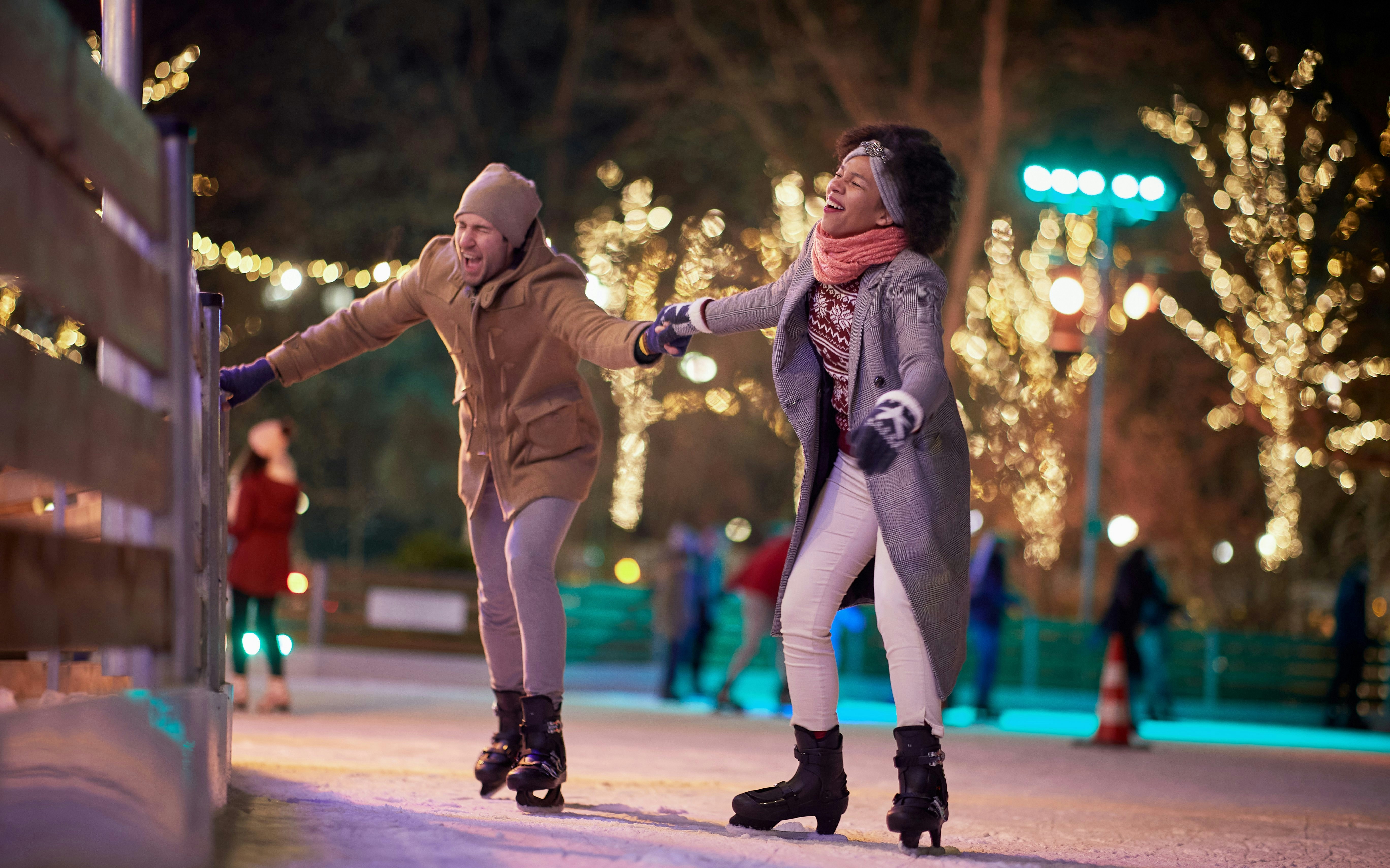 Couple ice skating at night, holding hands, surrounded by festive lights.