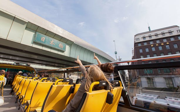 Tokyo hop-on hop-off bus passing under Nihonbashi bridge with passengers pointing.