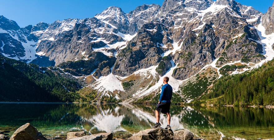 Man standing on rocks by Morskie Oko lake with snow-capped Tatra Mountains in Poland.