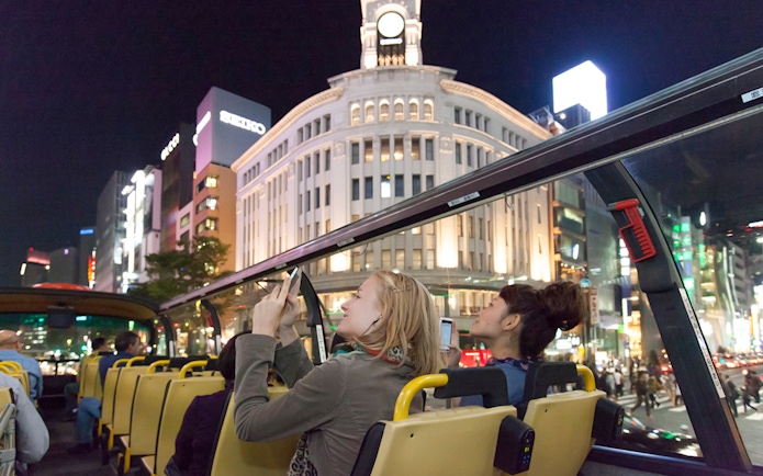 Open-top bus tour in Tokyo at night, passengers viewing illuminated Ginza buildings.