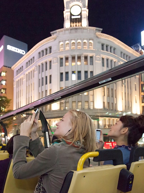 Open-top bus tour in Tokyo at night, passengers viewing illuminated Ginza buildings.