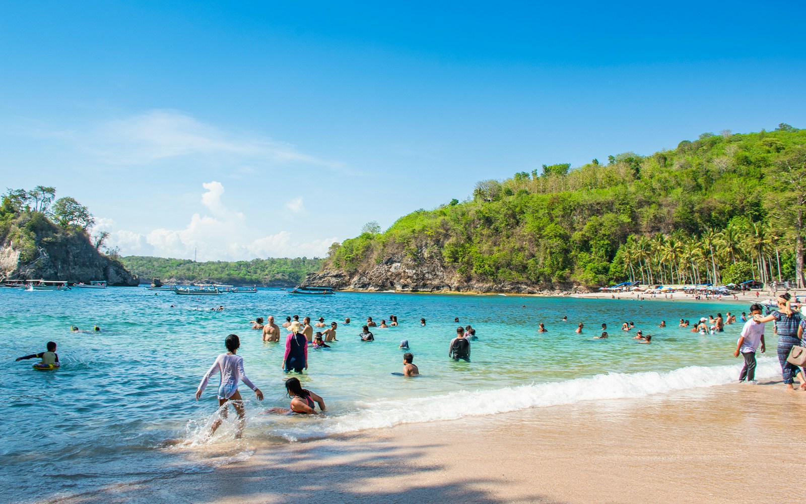 Tourists swimming at Crystal Bay Beach, West Nusa Penida Island, with lush hills in the background.