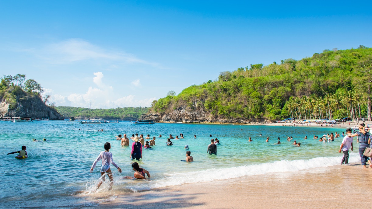Tourists swimming at Crystal Bay Beach, West Nusa Penida Island, with lush hills in the background.