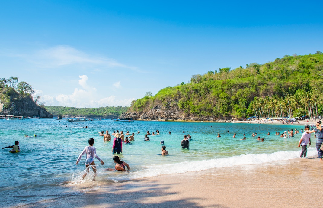 Tourists swimming at Crystal Bay Beach, West Nusa Penida Island, with lush hills in the background.