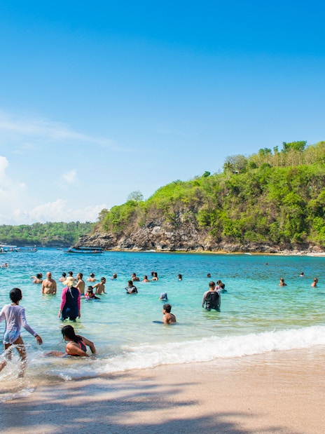 Tourists swimming at Crystal Bay Beach, West Nusa Penida Island, with lush hills in the background.