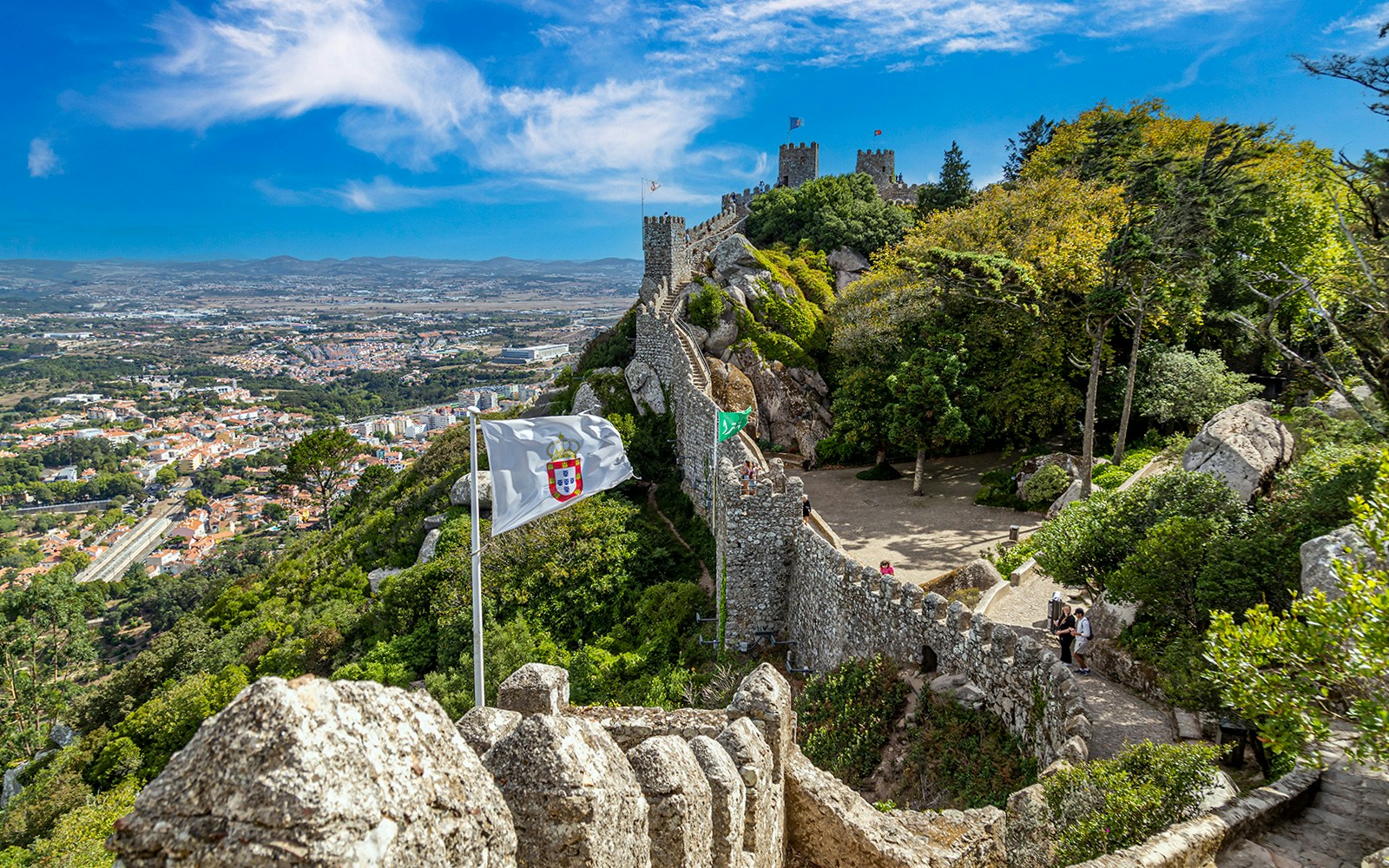 Ancient Moorish Castle walls overlooking Sintra city, Portugal, with lush greenery and flags.