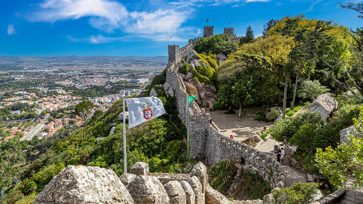 Ancient Moorish Castle walls overlooking Sintra city, Portugal, with lush greenery and flags.