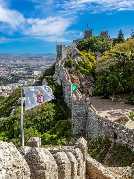 Ancient Moorish Castle walls overlooking Sintra city, Portugal, with lush greenery and flags.