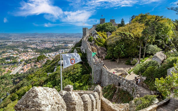 Ancient Moorish Castle walls overlooking Sintra city, Portugal, with lush greenery and flags.