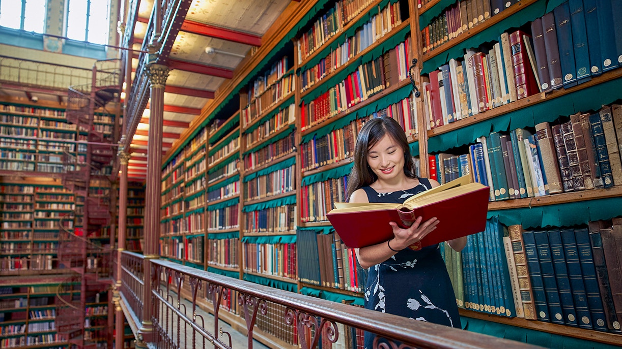 Person reading a large book in Cuypers Library at Rijksmuseum, surrounded by shelves of books.