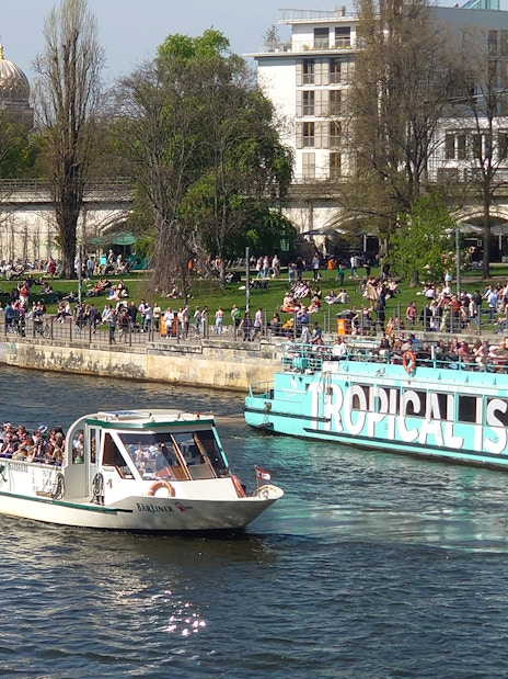 Sightseeing cruise boats on the Spree River in Berlin with people enjoying the riverside.