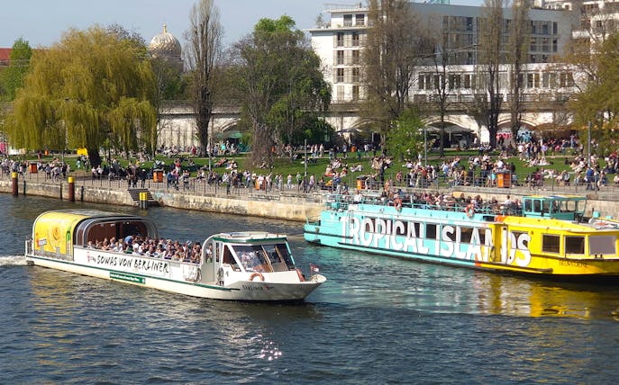 Sightseeing cruise boats on the Spree River in Berlin with people enjoying the riverside.