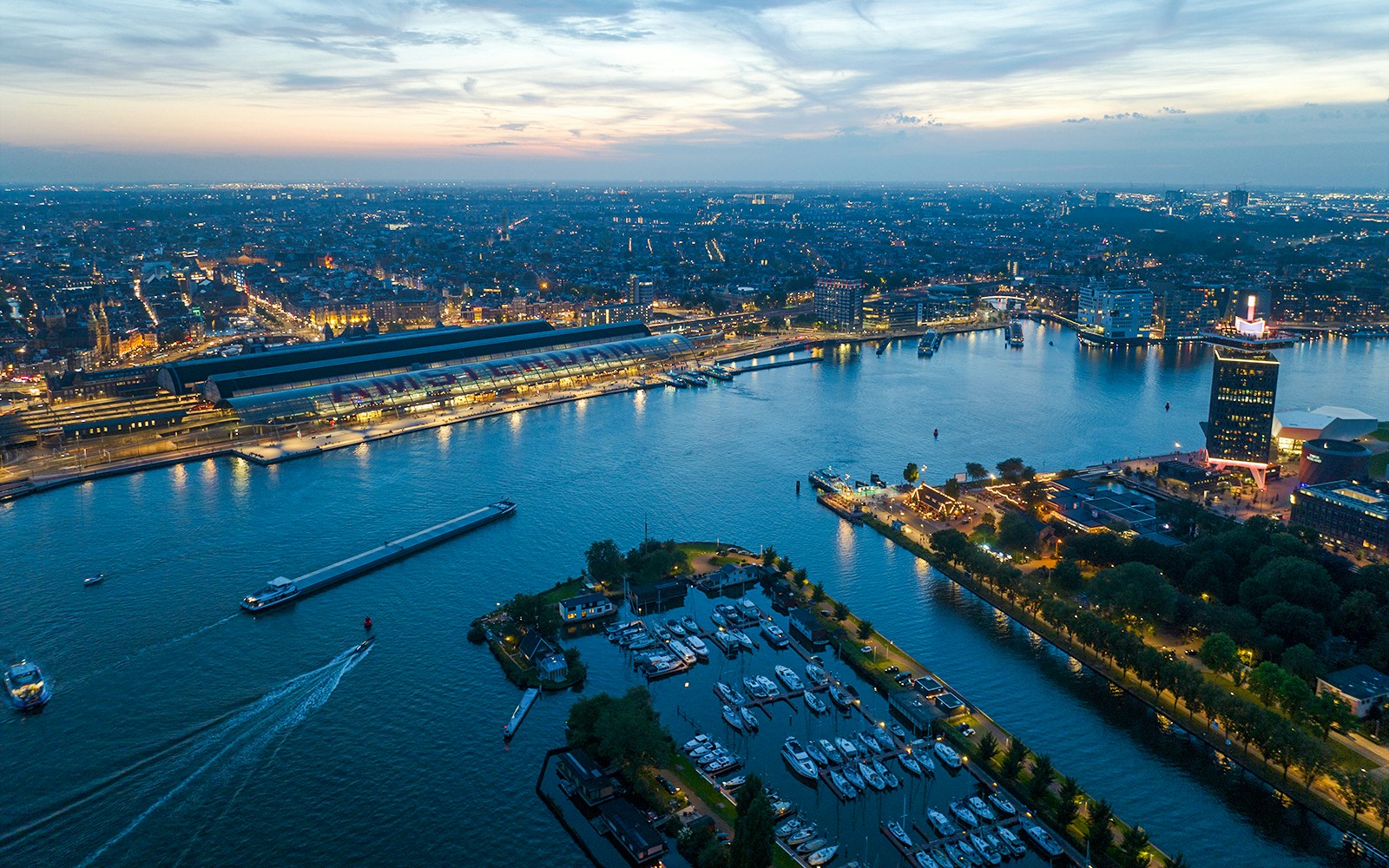 Aerial view of Amsterdam Centraal and Ij river at dusk, city lights reflecting on water.