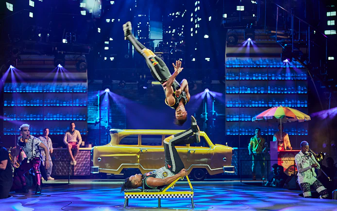 Acrobats performing flips on stage with a yellow taxi backdrop at Mad Apple show.