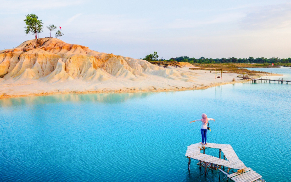 Person standing on a wooden pier over Blue Lake, Bintan, with sandy hills in the background.