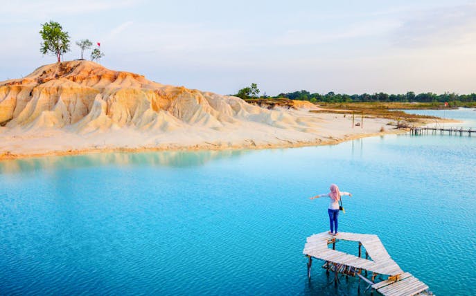 Person standing on a wooden pier over Blue Lake, Bintan, with sandy hills in the background.