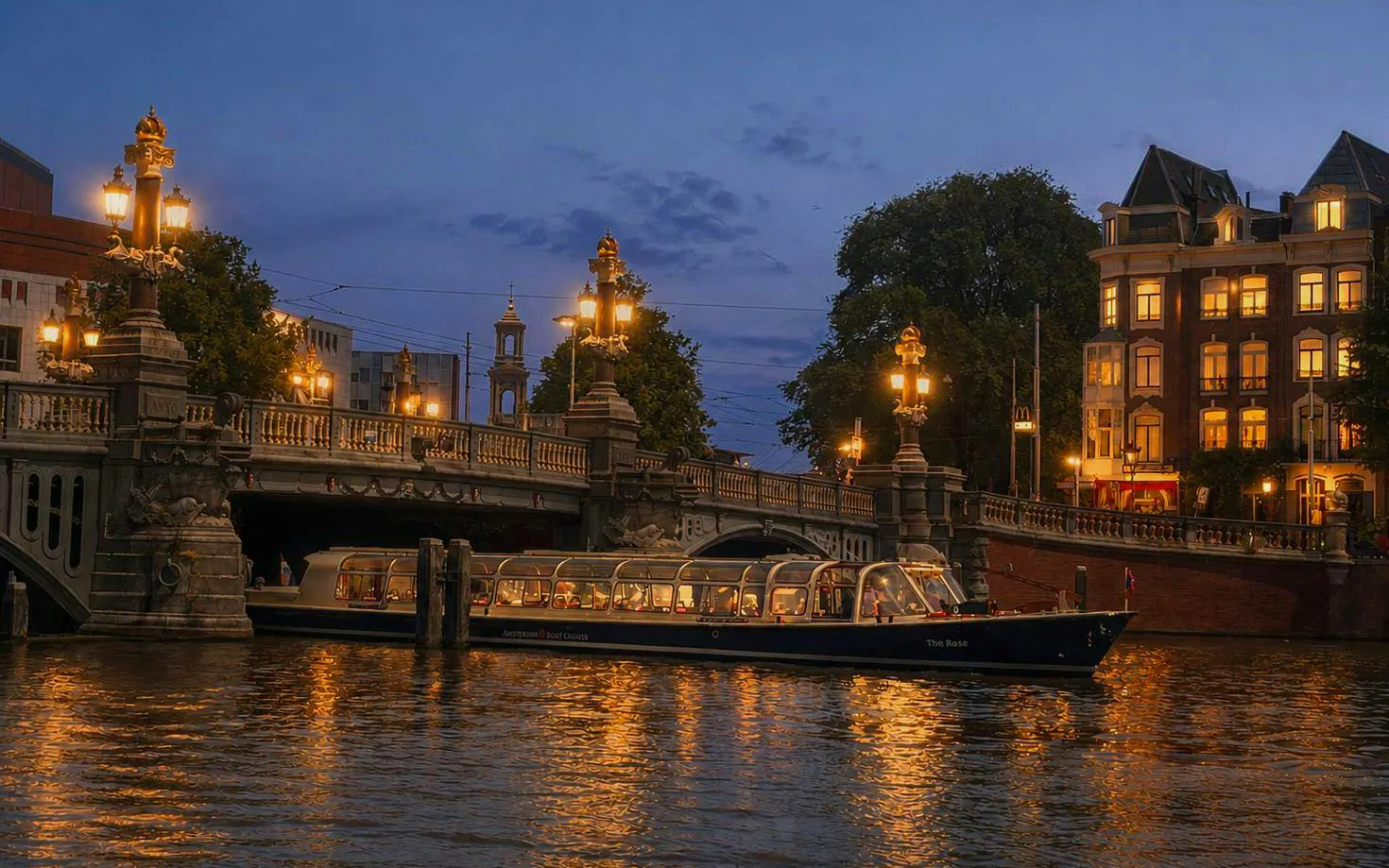 Amsterdam canal cruise boat passing under illuminated bridge at sunset.