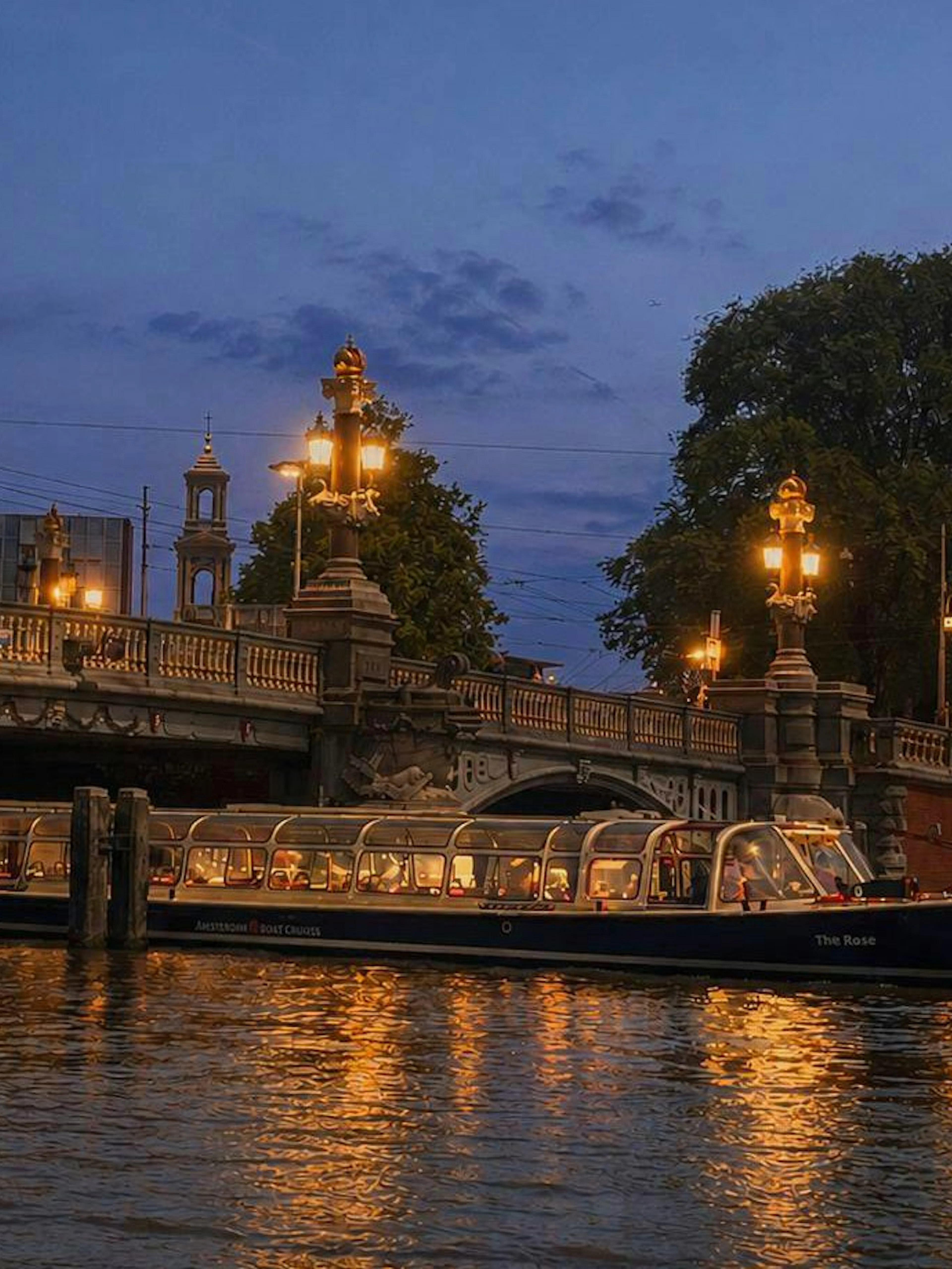Amsterdam canal cruise boat passing under illuminated bridge at sunset.