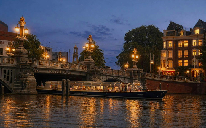 Amsterdam canal cruise boat passing under illuminated bridge at sunset.