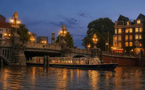 Amsterdam canal cruise boat passing under illuminated bridge at sunset.