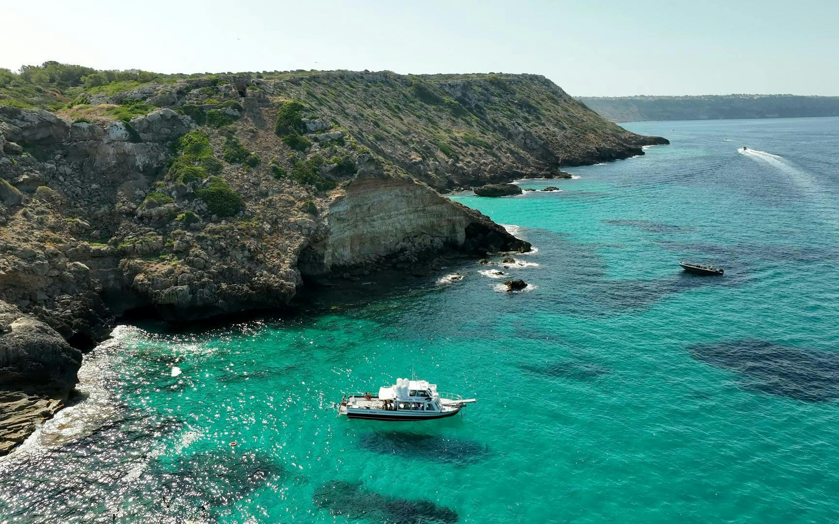 Catamaran sailing near rocky cliffs at Cala Vella, turquoise waters below.