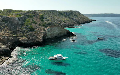 Catamaran sailing near rocky cliffs at Cala Vella, turquoise waters below.