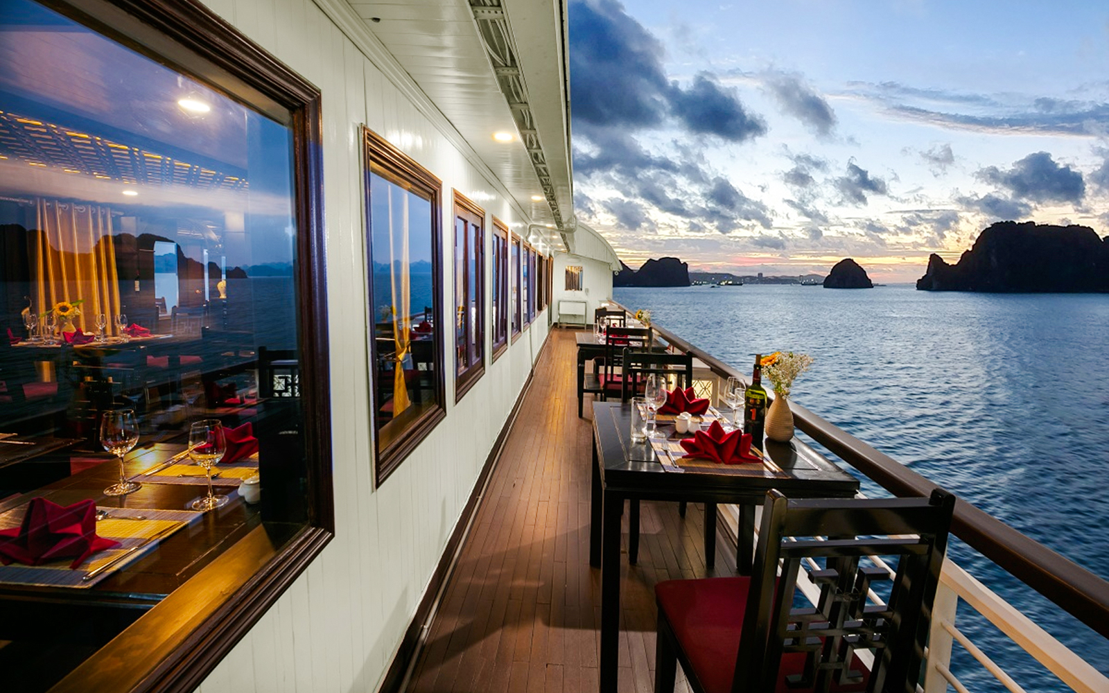 Dining tables on a cruise ship deck overlooking Ha Long Bay's limestone karsts at sunset.