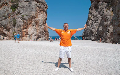 Man standing on a rocky beach between cliffs in Mallorca, with people and boats in the background.