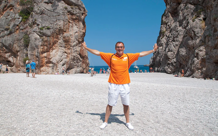 Man standing on a rocky beach between cliffs in Mallorca, with people and boats in the background.