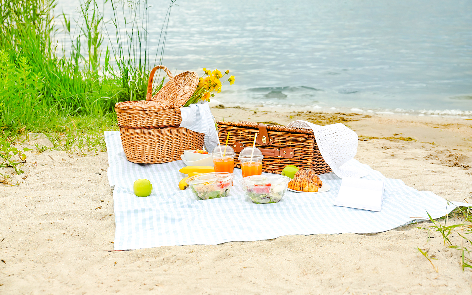 Beach picnic setup with baskets, salads, drinks, and fruit by the water.