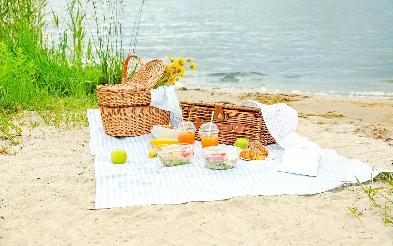 Beach picnic setup with baskets, salads, drinks, and fruit by the water.