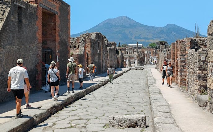 Visitors exploring ancient streets of Pompeii with Mount Vesuvius in the background.