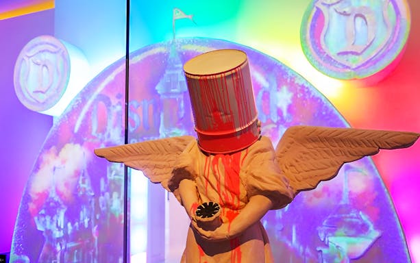 Banksy sculpture with paint bucket head in front of colorful backdrop.