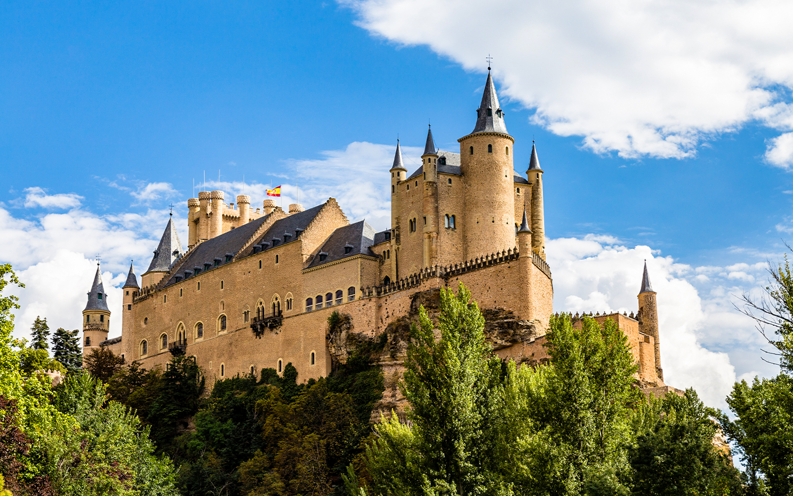 Alcazar of Segovia with towers and trees under a blue sky.