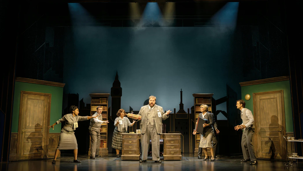 Actors performing a scene from "The Comedy About Spies" on stage with a cityscape backdrop.