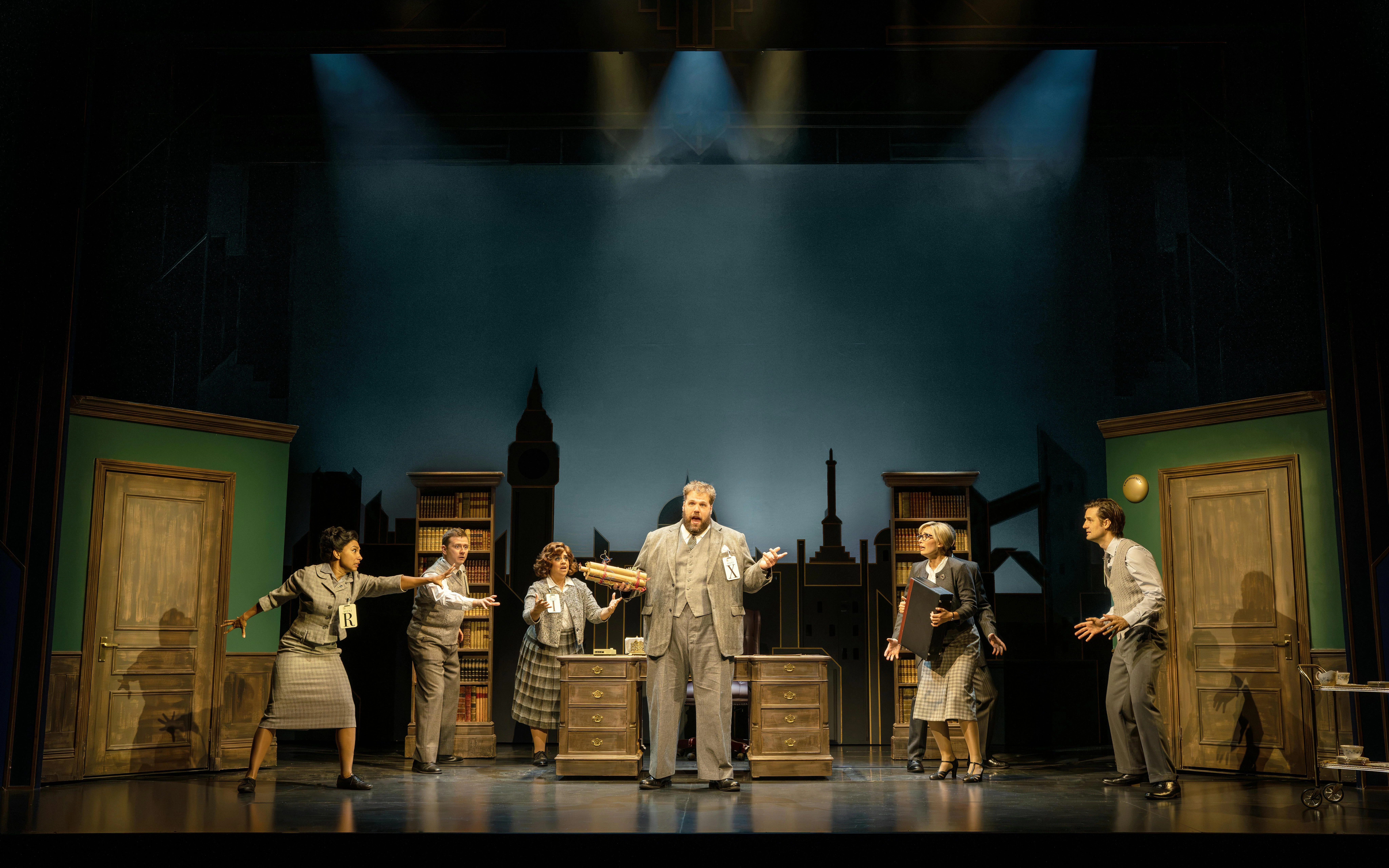 Actors performing a scene from "The Comedy About Spies" on stage with a cityscape backdrop.