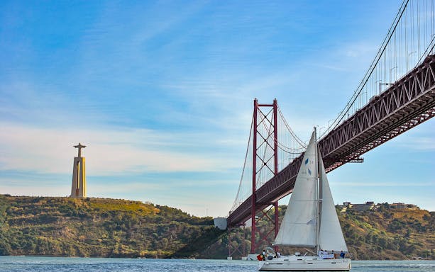 Sailboat on Tagus River near 25 de Abril Bridge and Cristo Rei statue in Lisbon.