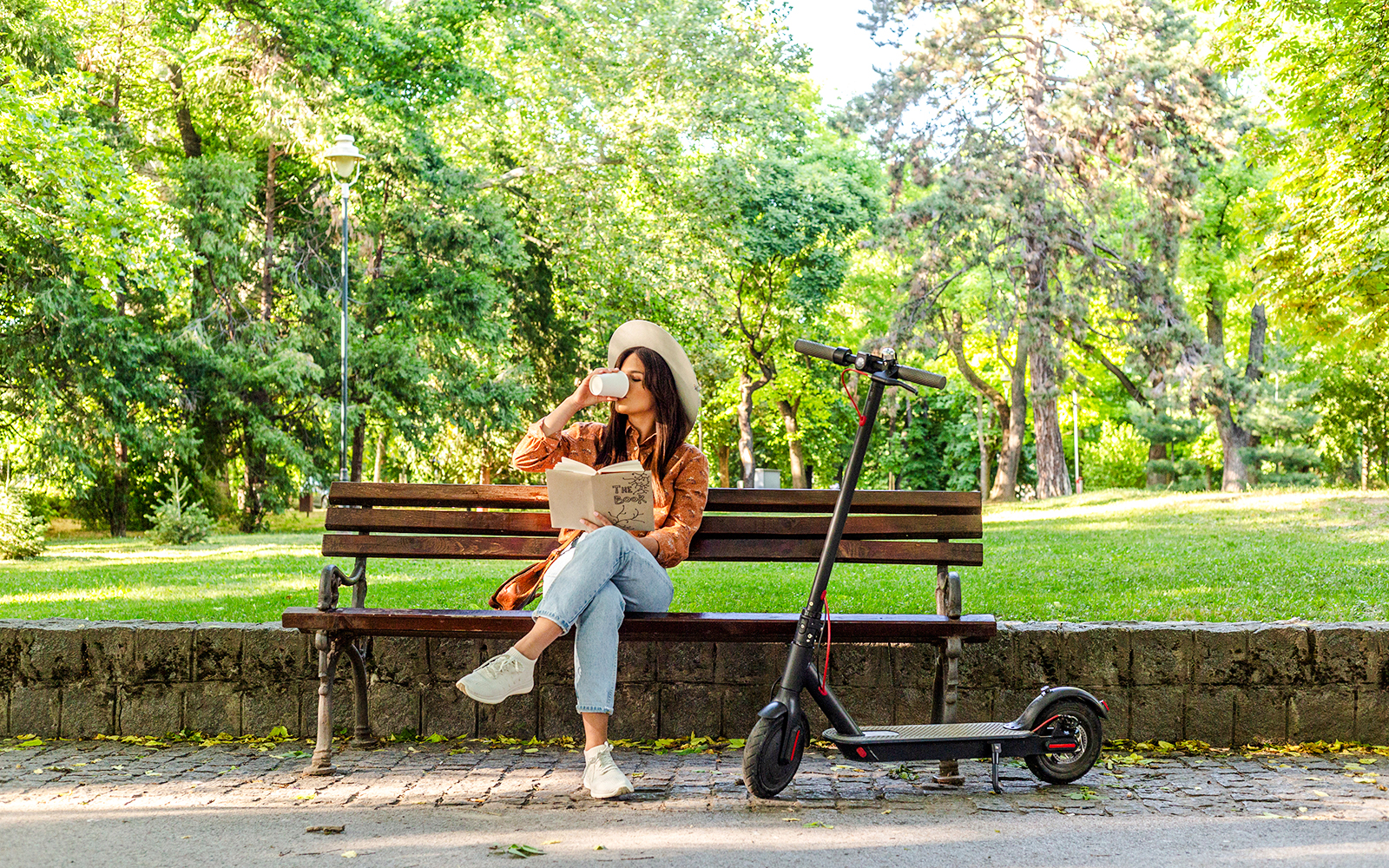 Person relaxing on a bench with a book and coffee, scooter nearby, in Jardin des Bassins.